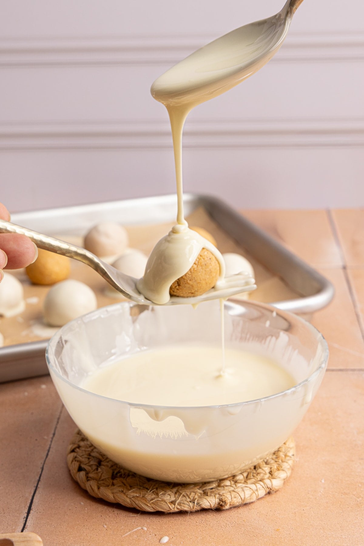 A fork holds a ball over a bowl of melted white chocolate and a spoon drizzles the chocolate over the truffle.