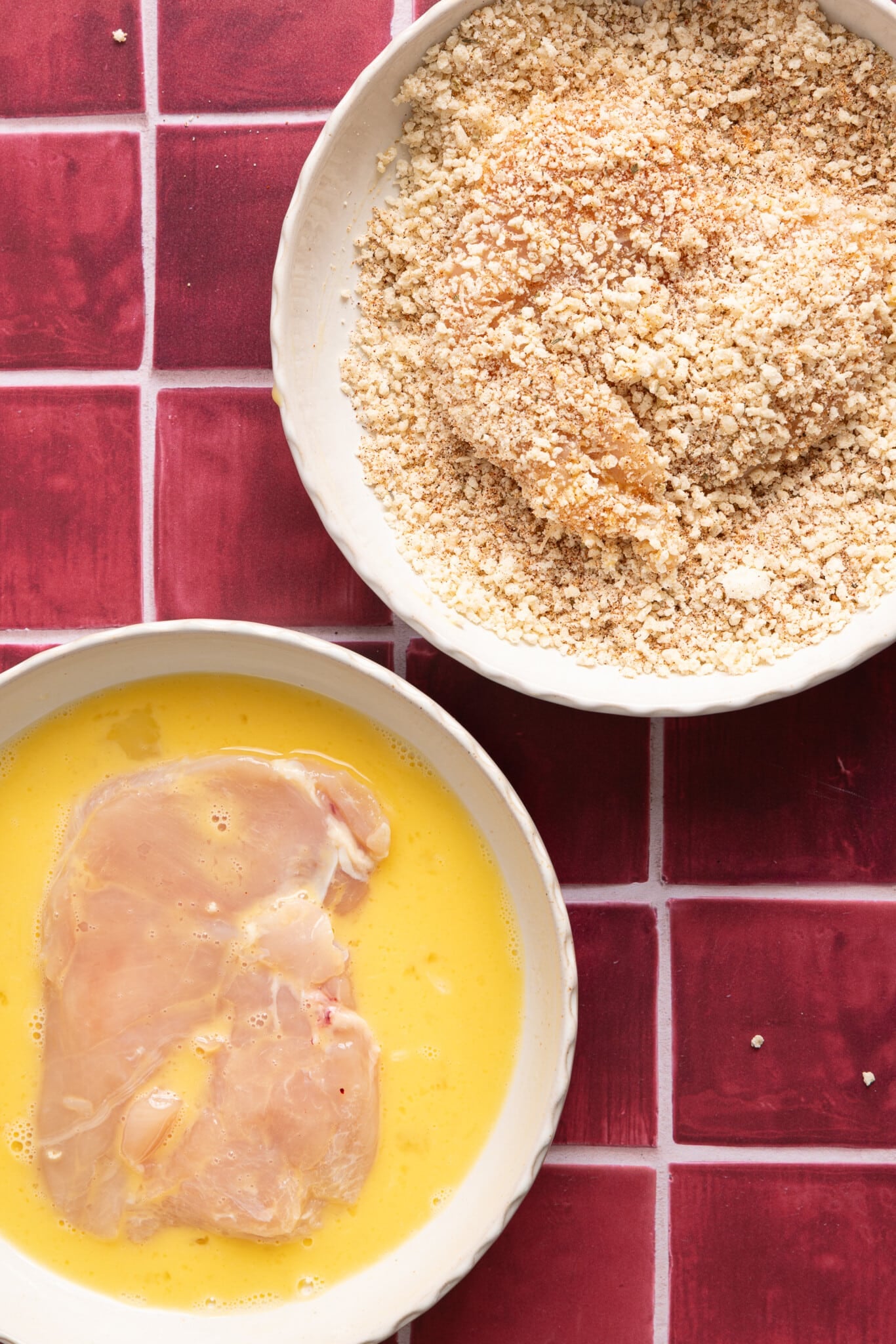 A chicken cutlet rests in the bowl of whisked egg and a second chicken cutlets lays in the bowl of breadcrumb topping.